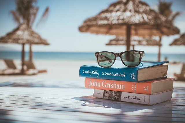 Kôpka kníh s okuliarmi na terase, ktorá je na pláži, v pozadí je slnečník.jpg